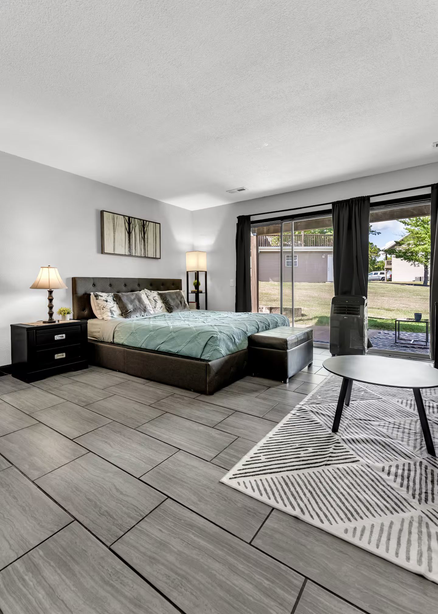 A modern bedroom with a large bed, a nightstand, a bedside table, a lamp, a chair, and a view of the outdoors.