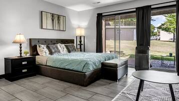 A modern bedroom with a large bed, a nightstand, a bedside table, a lamp, a chair, and a view of the outdoors.