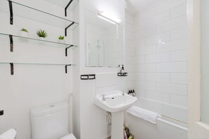 Ein Badezimmer mit Toilette, Waschbecken, Spiegel und Dusche. Regale mit Pflanzen und Pflegeprodukten sind vorhanden.