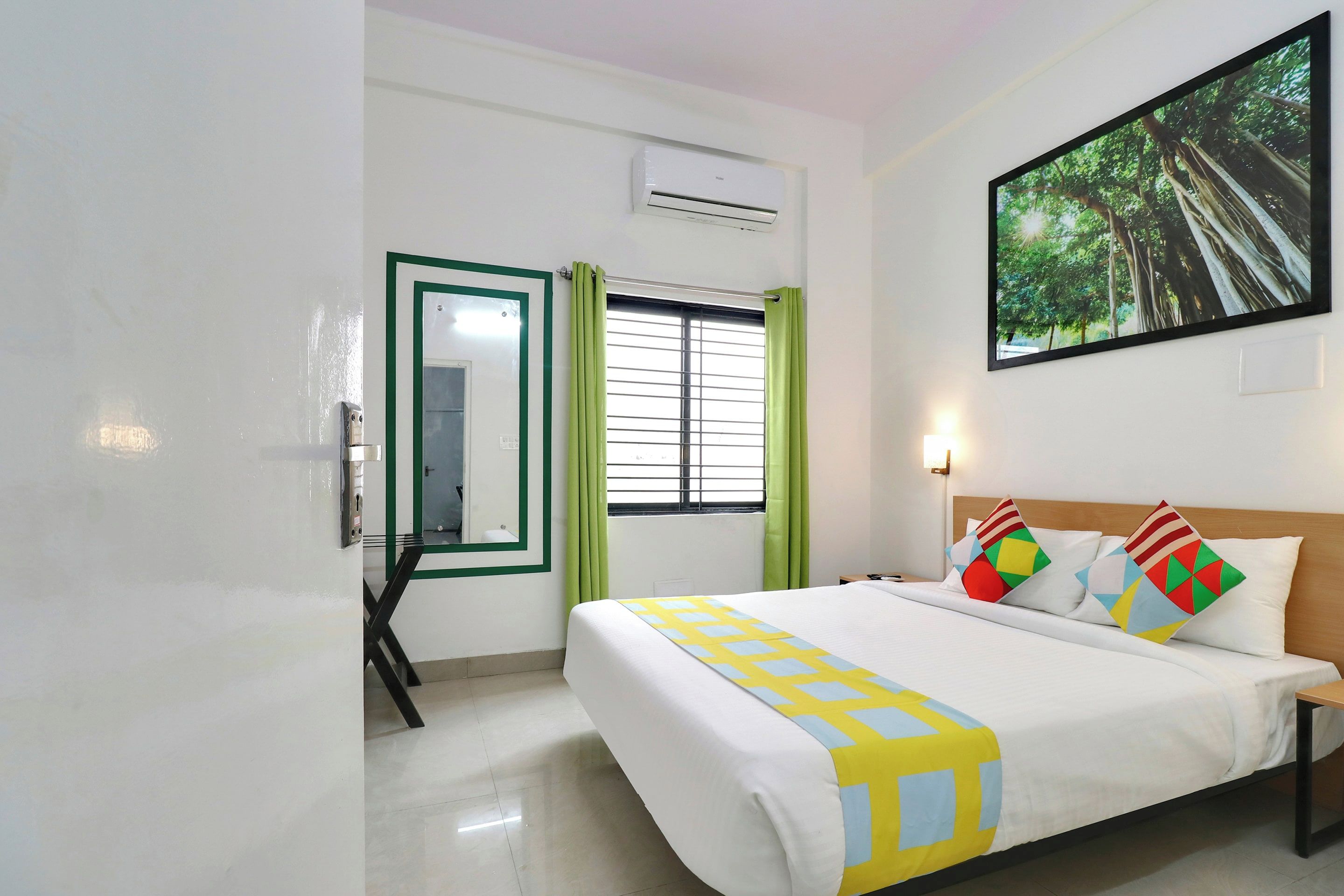 A hotel room with a bed, a window with blinds, a painting on the wall, and an air conditioning unit.