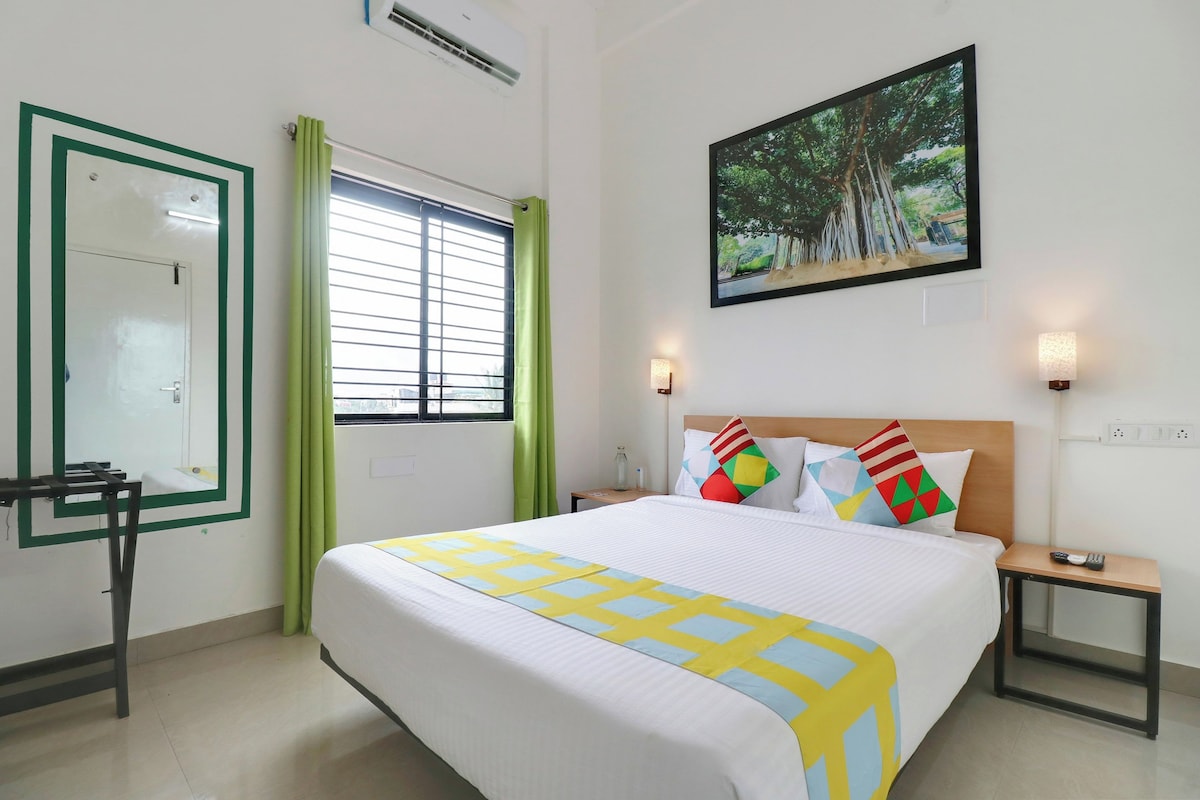 A hotel room with a bed, a nightstand, a wall-mounted TV, a window with curtains, and a painting of trees.