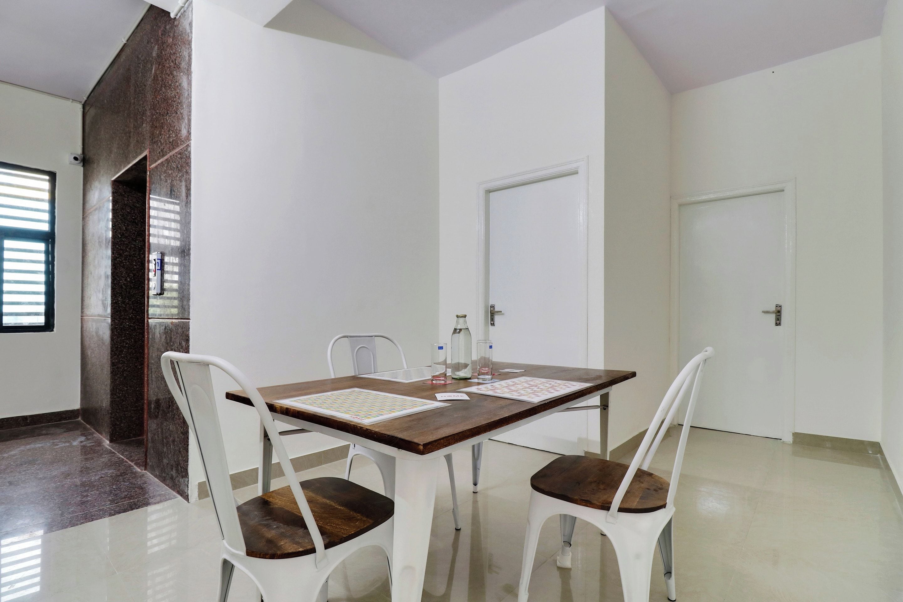 Un área de comedor con una mesa de madera y sillas blancas, una puerta y un ventanal con persianas.
