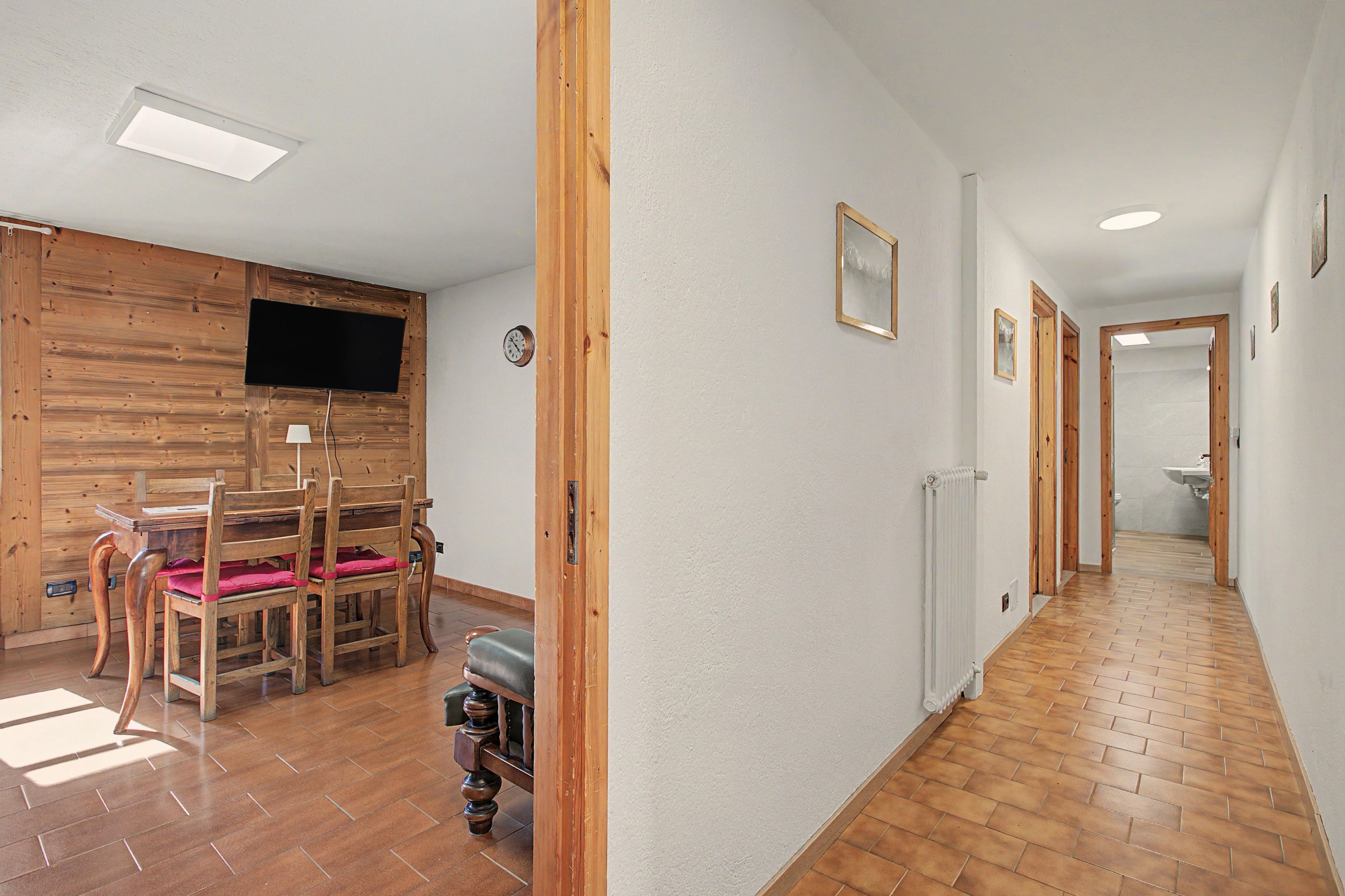 A hallway with wooden flooring, a dining area with a table and chairs, a television mounted on a wooden wall, and a radiator along the wall.