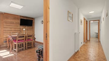 A hallway with wooden flooring, a dining area with a table and chairs, a television mounted on a wooden wall, and a radiator along the wall.