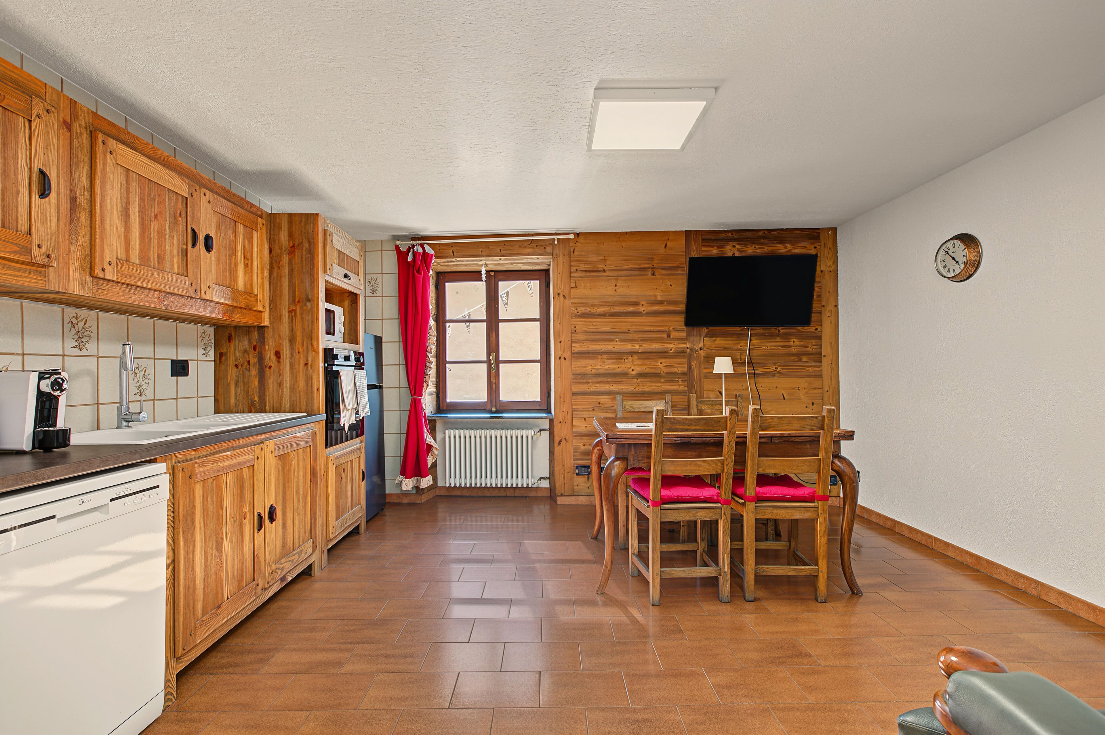 Een keuken met houten keukenkastjes, een eettafel met stoelen, een raam met gordijnen en een klok aan de muur.