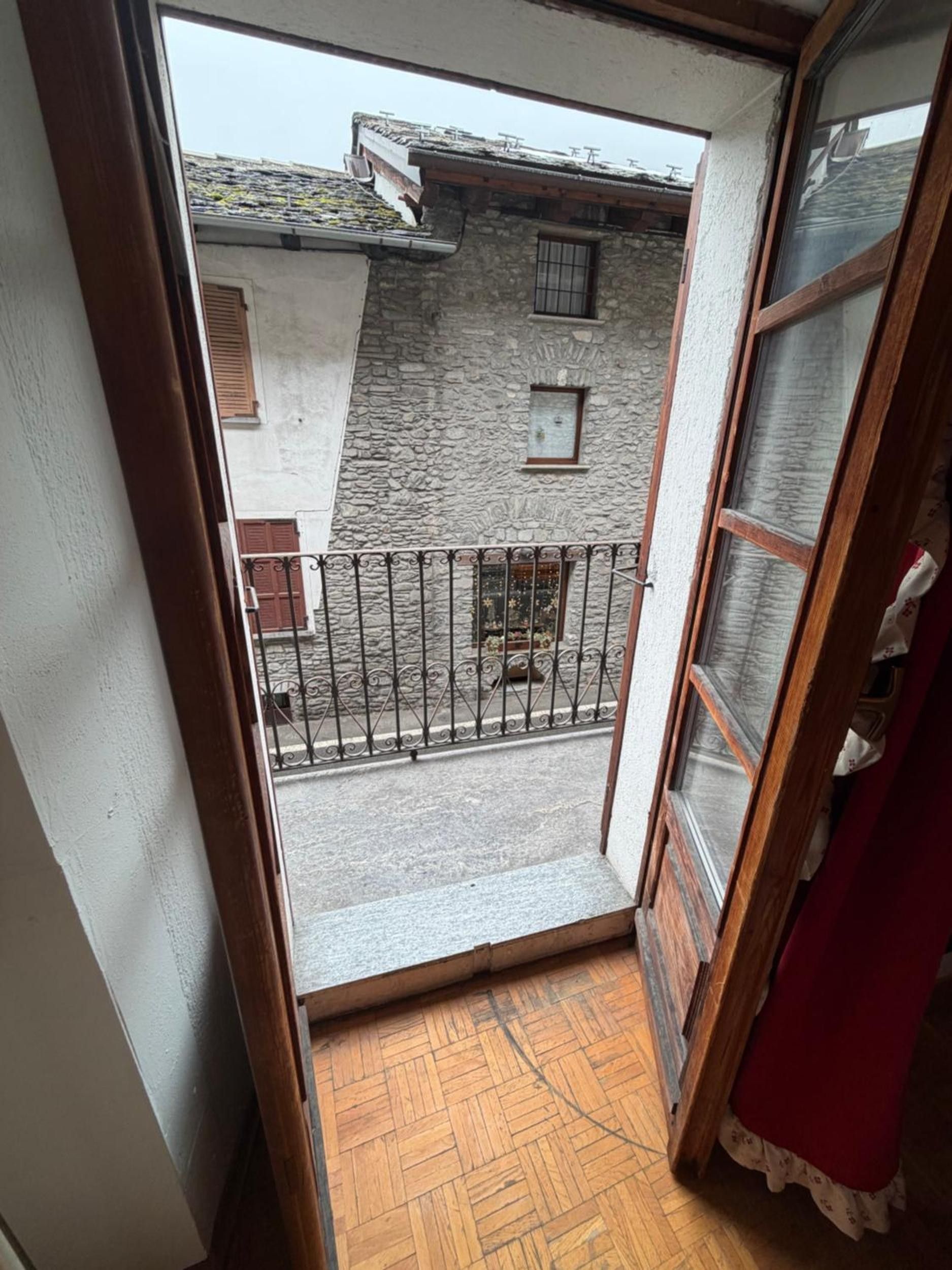 A narrow balcony with a wooden floor, a metal railing, and a view of a stone building with a window and shutters.