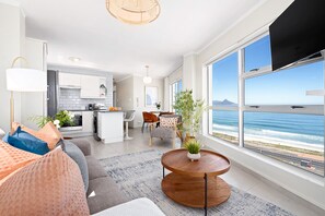 A modern living room with a large window overlooking the ocean, a kitchen in the background, and a dining area with a view. - The Bay A801 (Cape Town)