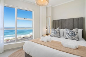Ein Schlafzimmer mit großem Fenster und Blick auf den Strand, einem Bett mit weißen Leinen und einem grau gepolsterten Kopfteil.