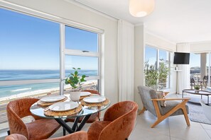 Ein modernes Wohnzimmer mit großen Fenstern mit Blick auf einen Strand, ein Essbereich mit Tisch für vier Personen und eine Sitzecke mit Fernseher und Pflanzen.