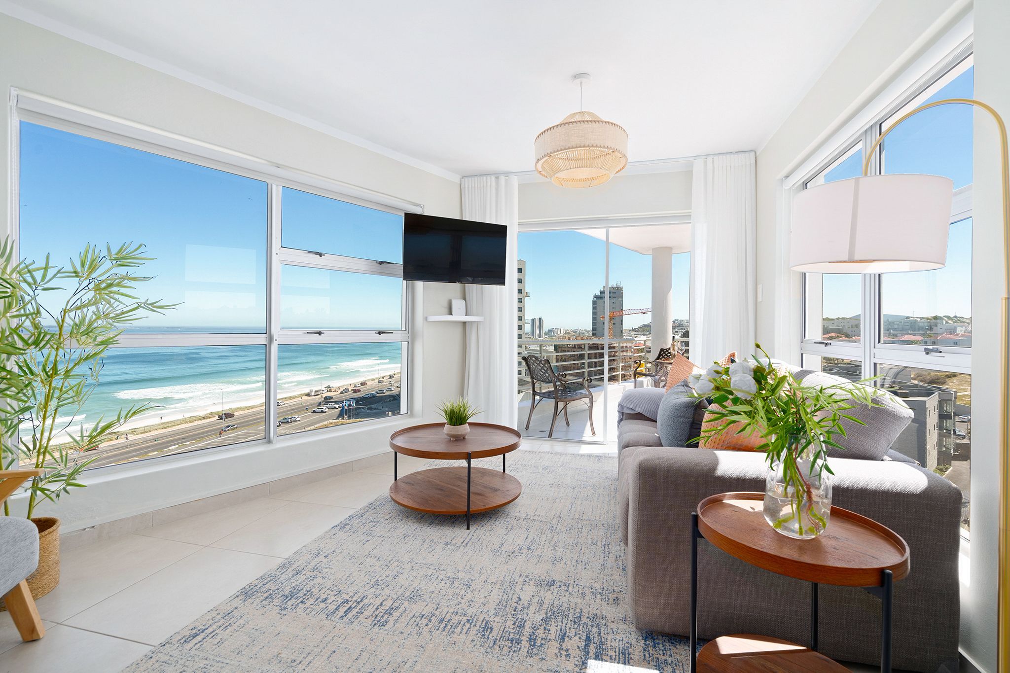 A modern living room with a large window overlooking a beach and cityscape, furnished with a sofa, coffee table, and a TV.