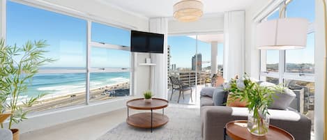 A modern living room with a large window overlooking a beach and cityscape, furnished with a sofa, coffee table, and a TV.