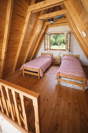 Un pequeño dormitorio en el ático, de madera, con dos camas, una ventana y techo de madera.