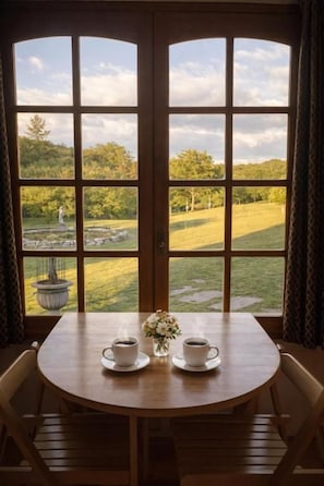 Una habitación con un ventanal grande, una mesa redonda de madera con dos tazas de café y un jarrón con flores.