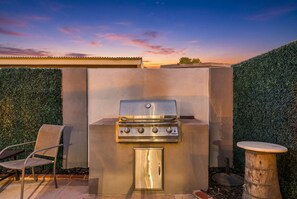 An outdoor grill with a stainless steel finish, a chair, and a small table, set against a backdrop of a hedge and a sunset sky. - 3BR Scottsdale Retreat, Pool, Near Old Town (Scottsdale)
