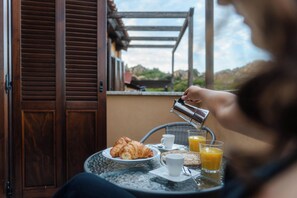 Eine Person gießt ein Getränk in ein Glas auf einem Tisch mit Croissants und einer Tasse, draußen mit einer Holzkonstruktion und Blick auf Grünflächen.