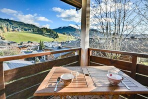 Een houten tafel met twee kommen en lepels op een balkon met uitzicht op een dorp en bergen.