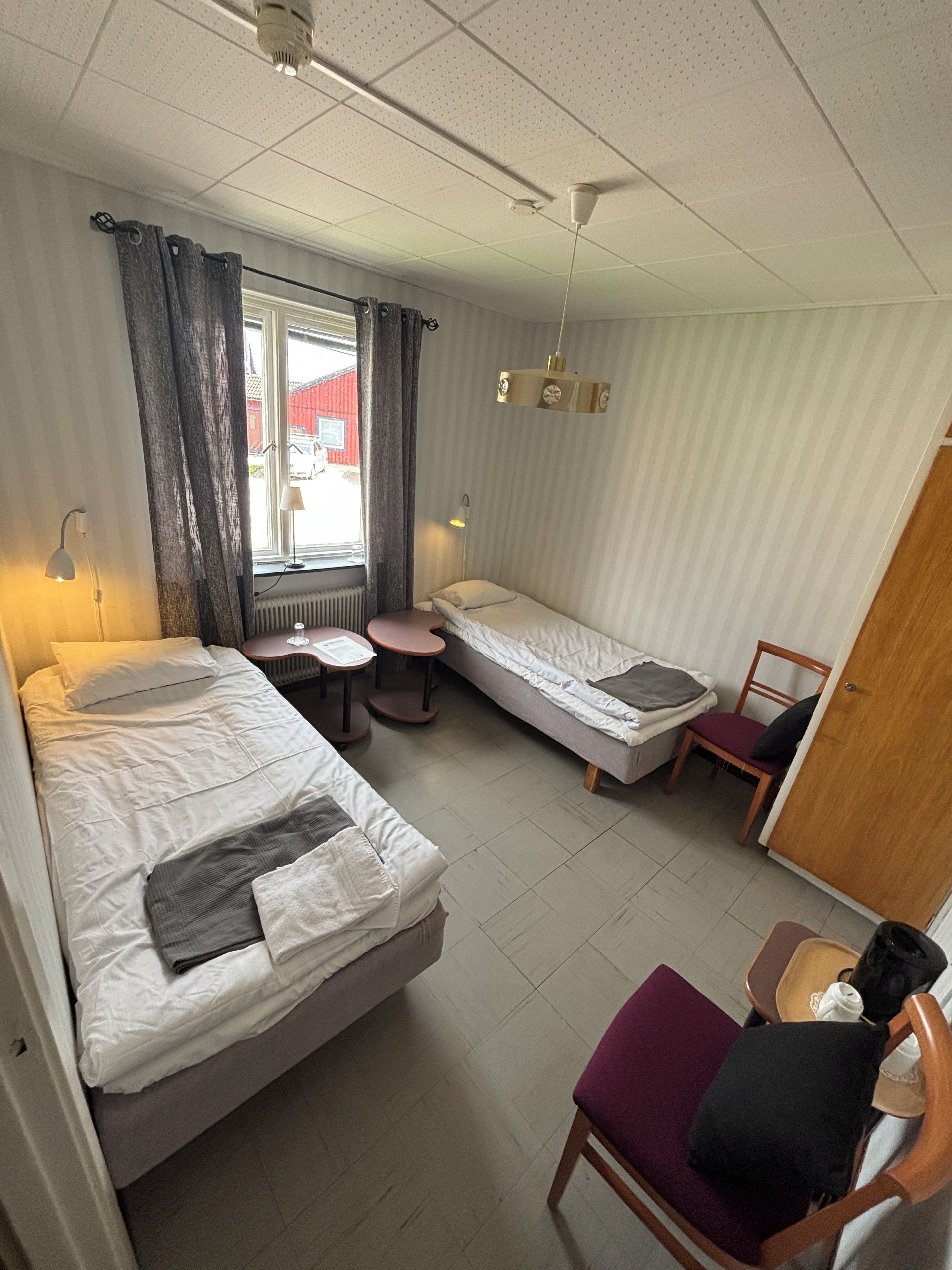 A compact hotel room with two single beds, a small table, a chair, and a window with curtains.