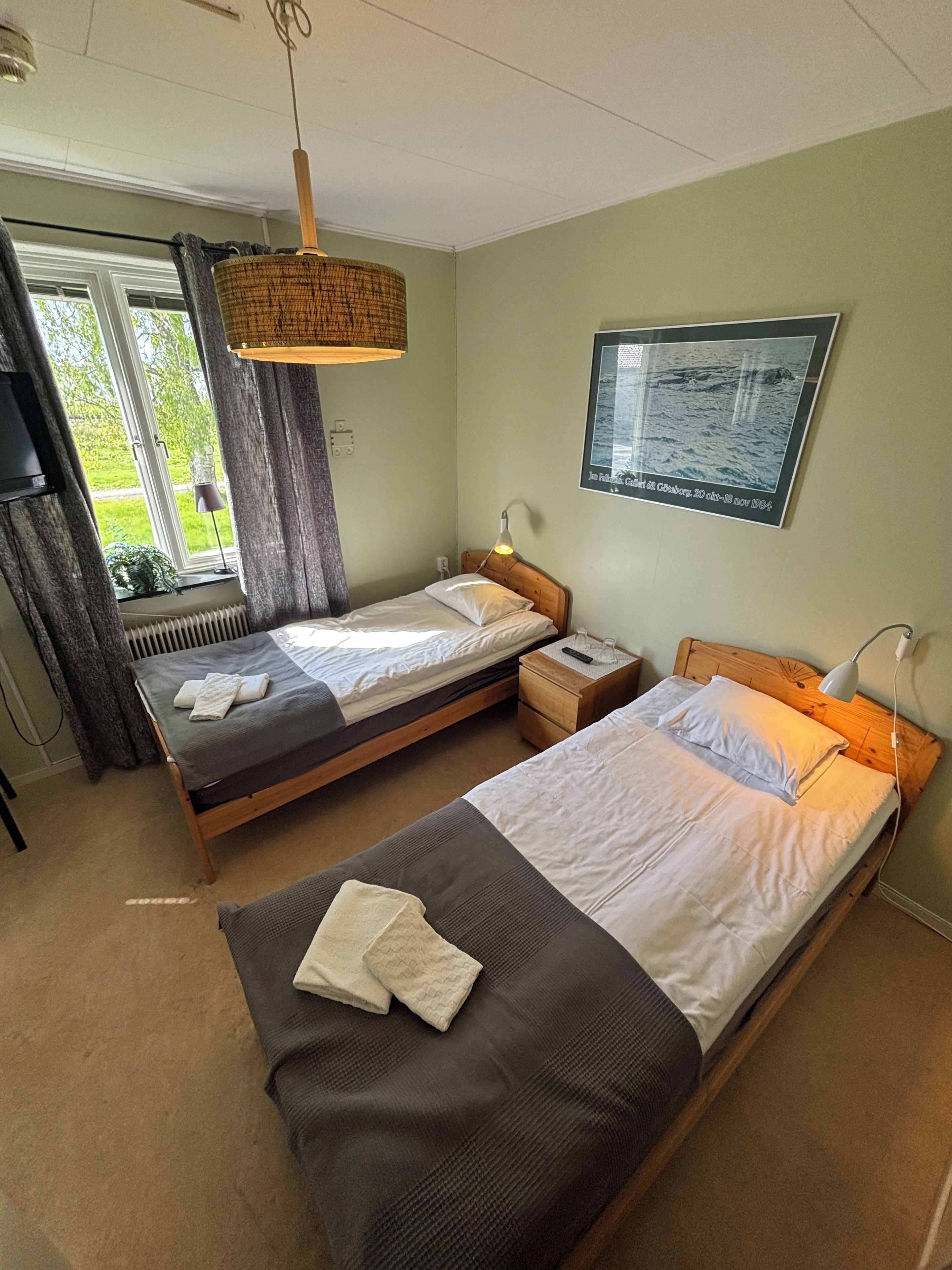 A room with two beds, a window with curtains, a wall-mounted picture, and a bedside lamp.