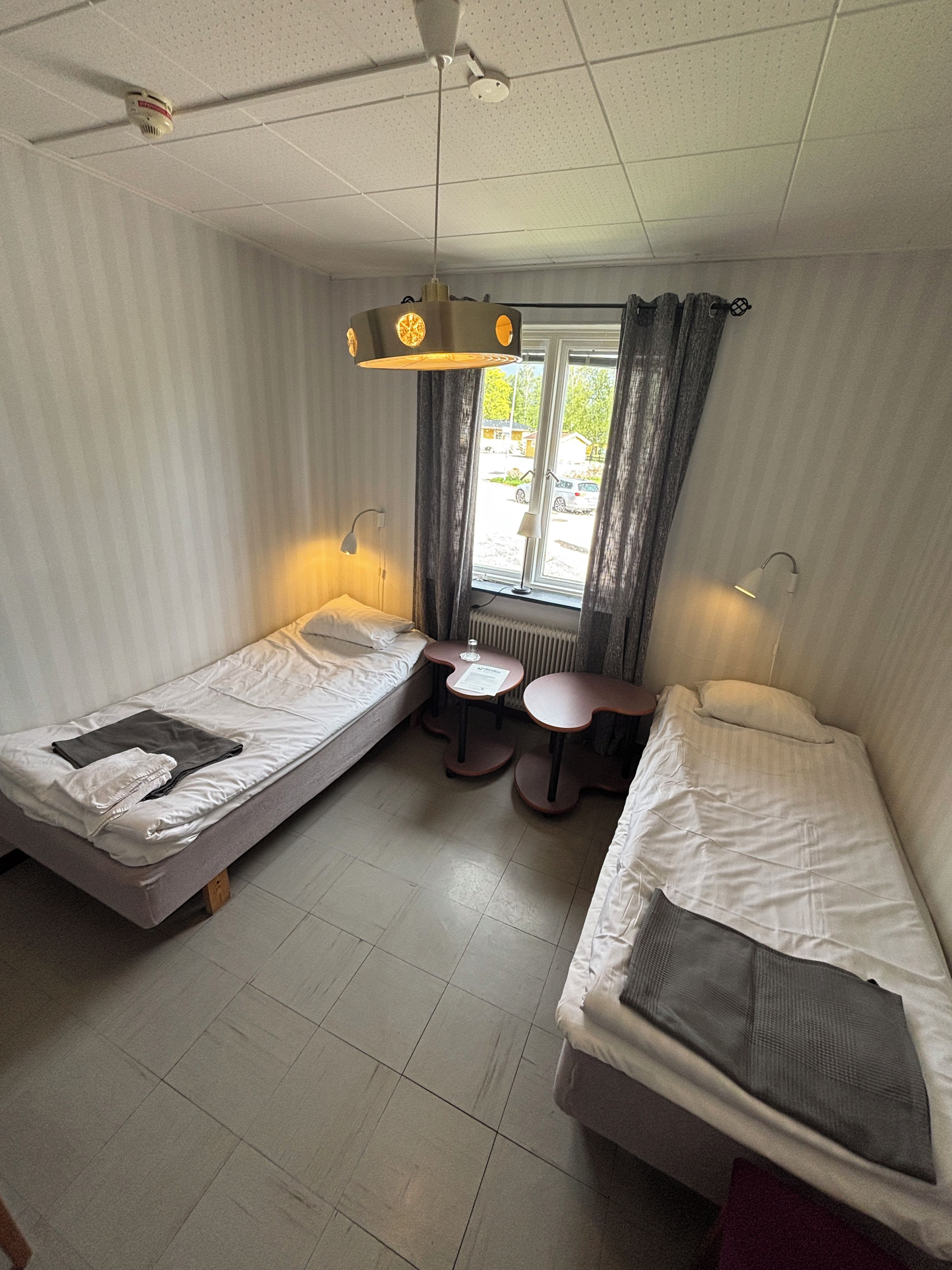 Una habitación con dos camas individuales, una mesita pequeña, proyector en el techo y ventana con cortinas.