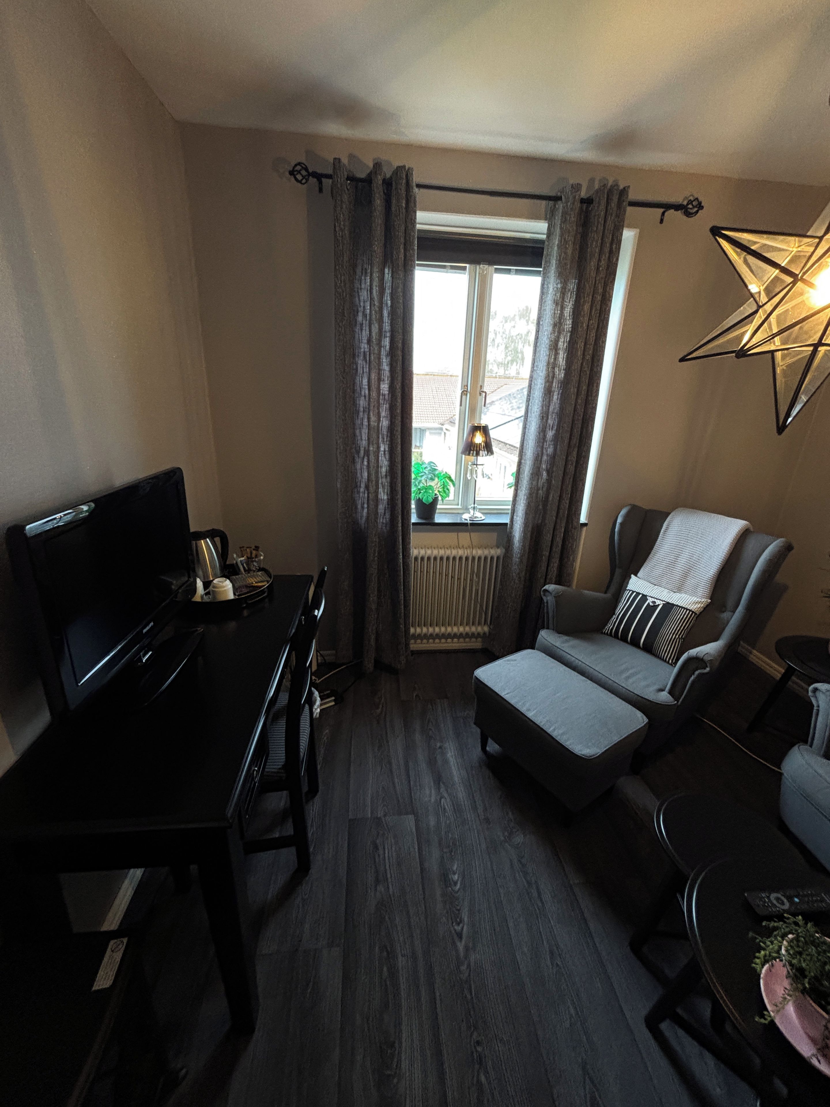 Ein kompakter Wohnraum mit dunklem Holzboden, einem schwarzen Schreibtisch mit Fernseher, einem grauen Sofa und einer sternförmigen Leuchte.
