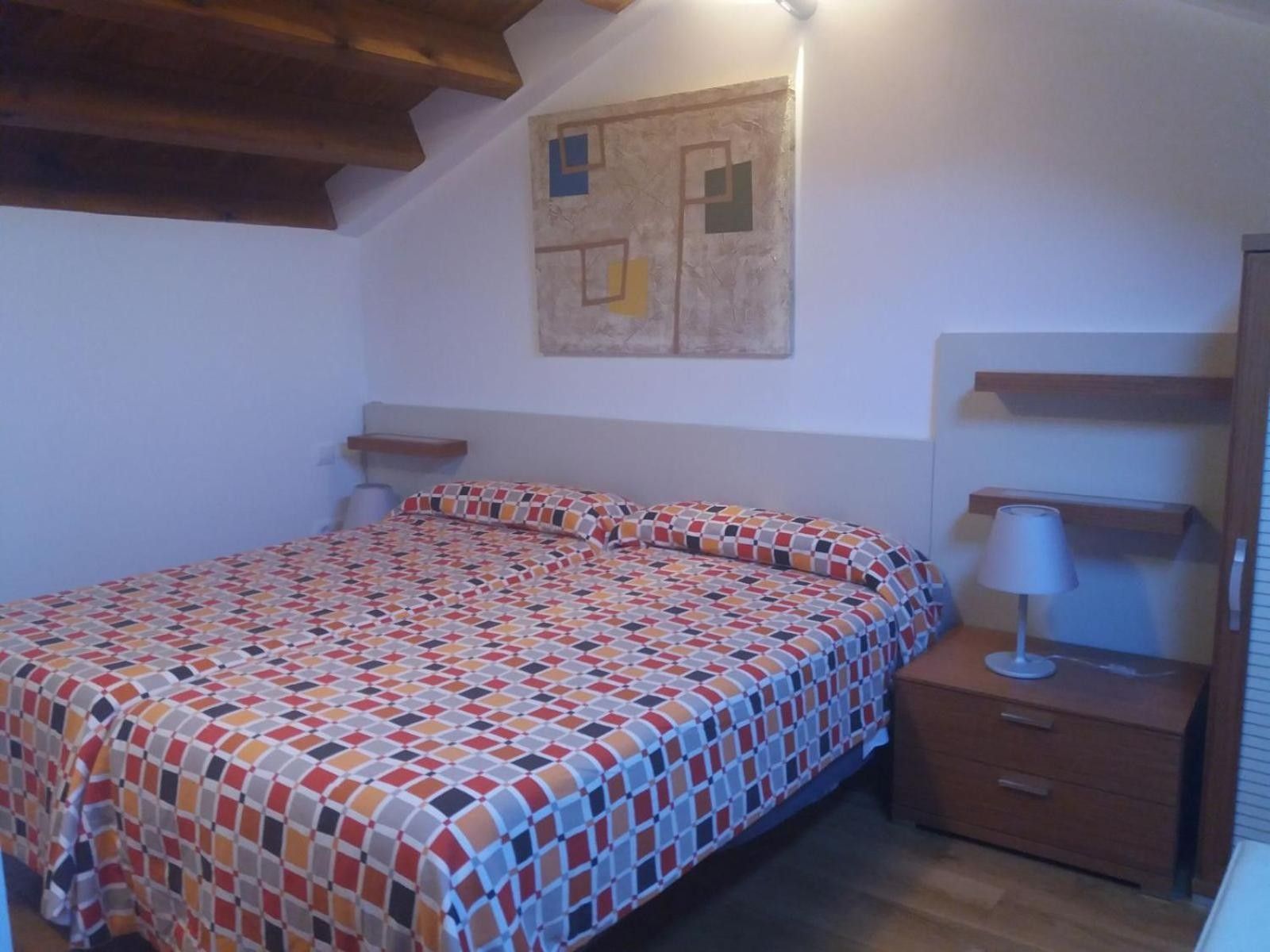 A bedroom with a patterned bedspread, a nightstand with a lamp, and a wall-mounted shelf.