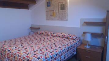A bedroom with a patterned bedspread, a nightstand with a lamp, and a wall-mounted shelf.