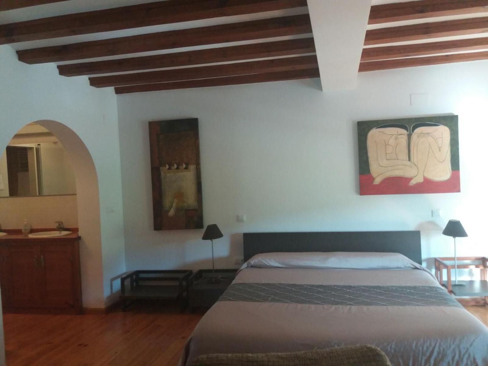 A bedroom with a wooden ceiling, a bed with a patterned bedspread, a nightstand, a mirror, and a painting on the wall.
