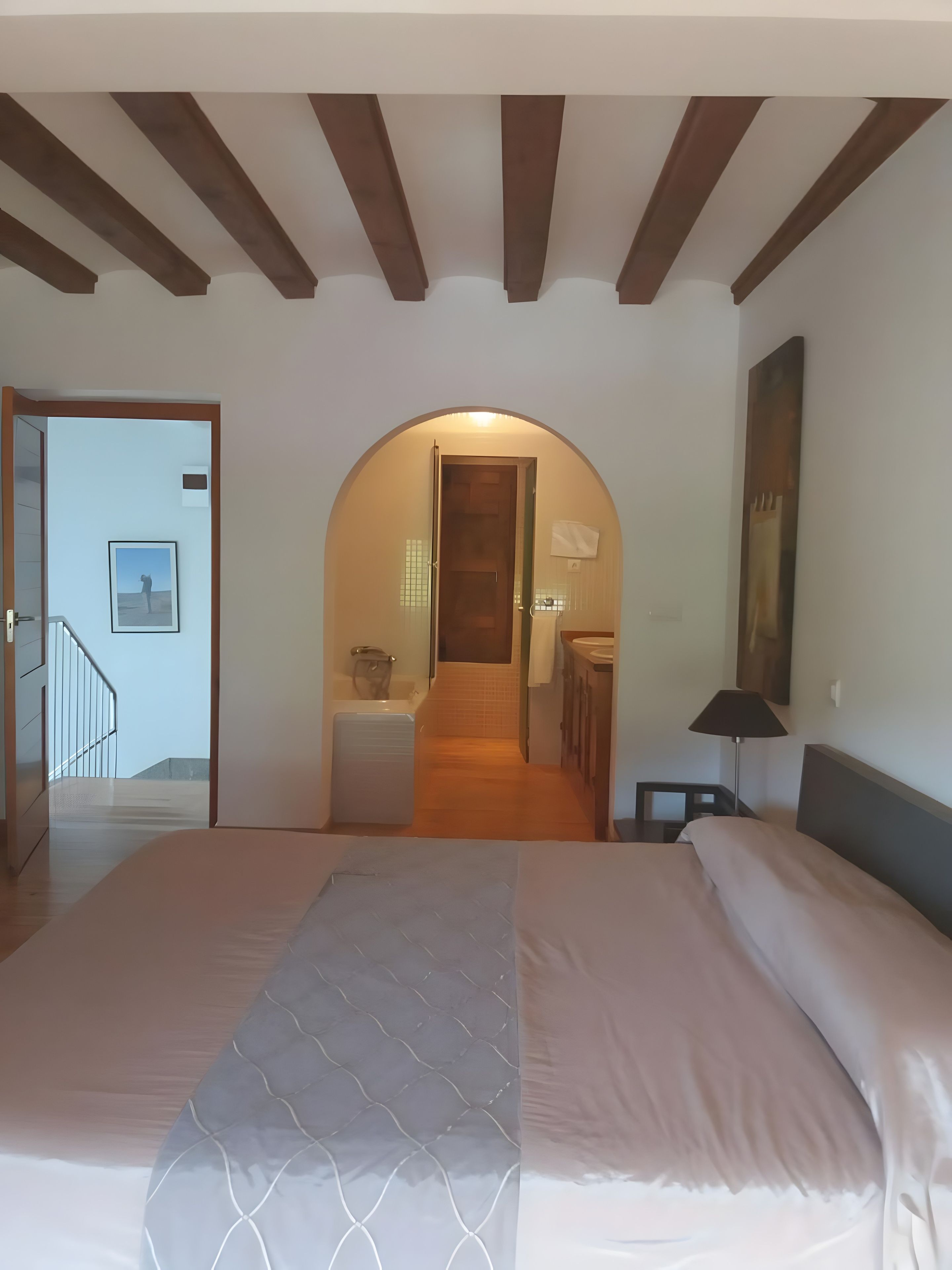 Een slaapkamer met een groot bed, een houten plafond, een badkamer met douche en een gang die naar een andere kamer leidt.