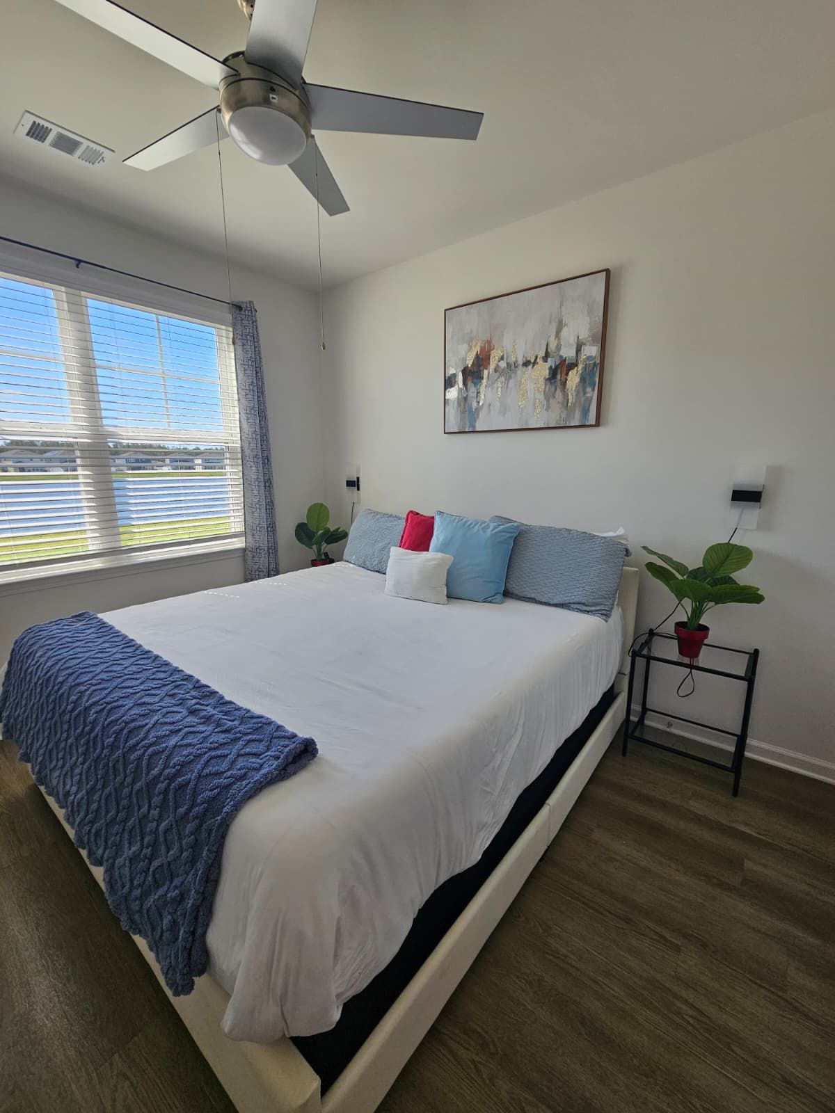 A bedroom with a bed, a ceiling fan, a window with blinds, a painting, and a bedside plant stand.