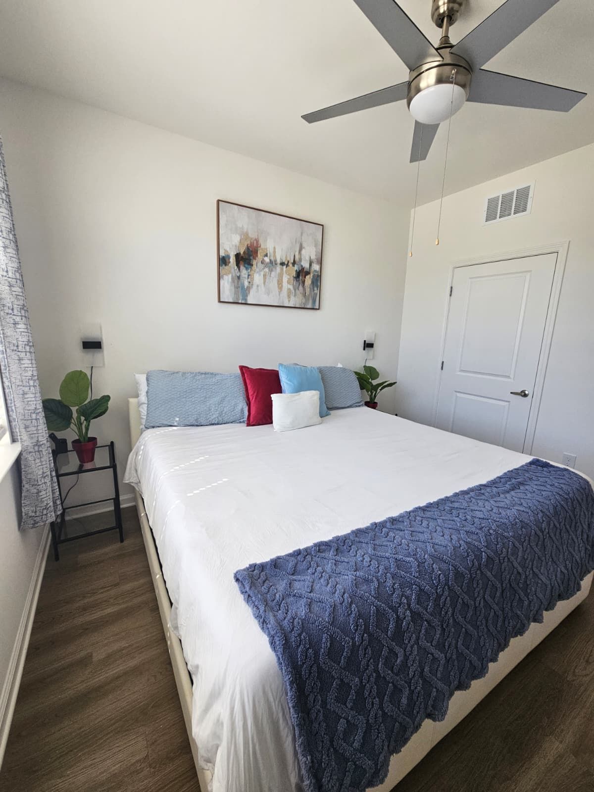 A bedroom with a bed, a ceiling fan, a door, a window with curtains, and a wall-mounted abstract painting.