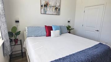 A bedroom with a bed, a ceiling fan, a door, a window with curtains, and a wall-mounted abstract painting.