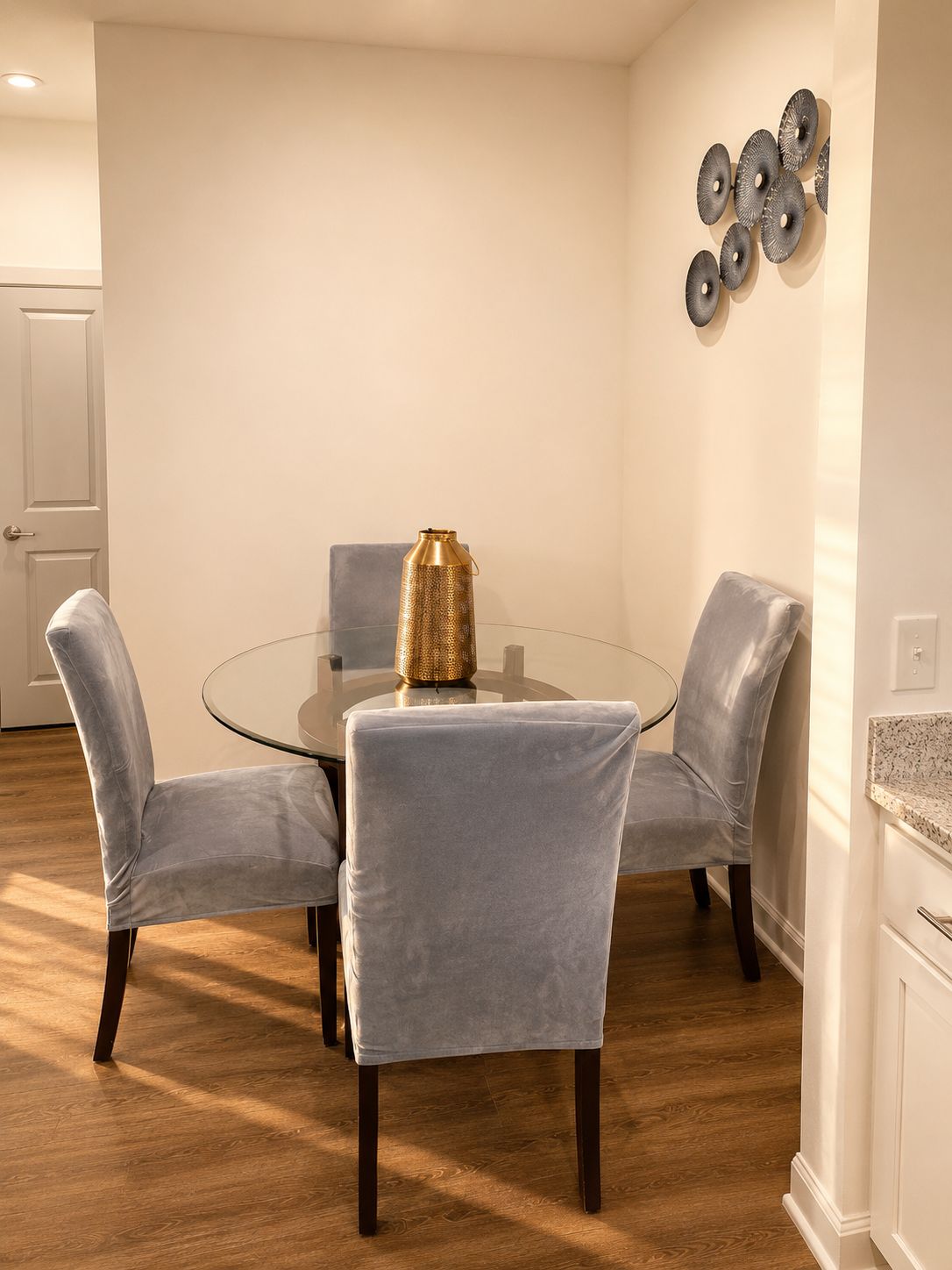 A dining area with a round glass table, four grey upholstered chairs, a gold textured vase, and a wall-mounted circular decoration.
