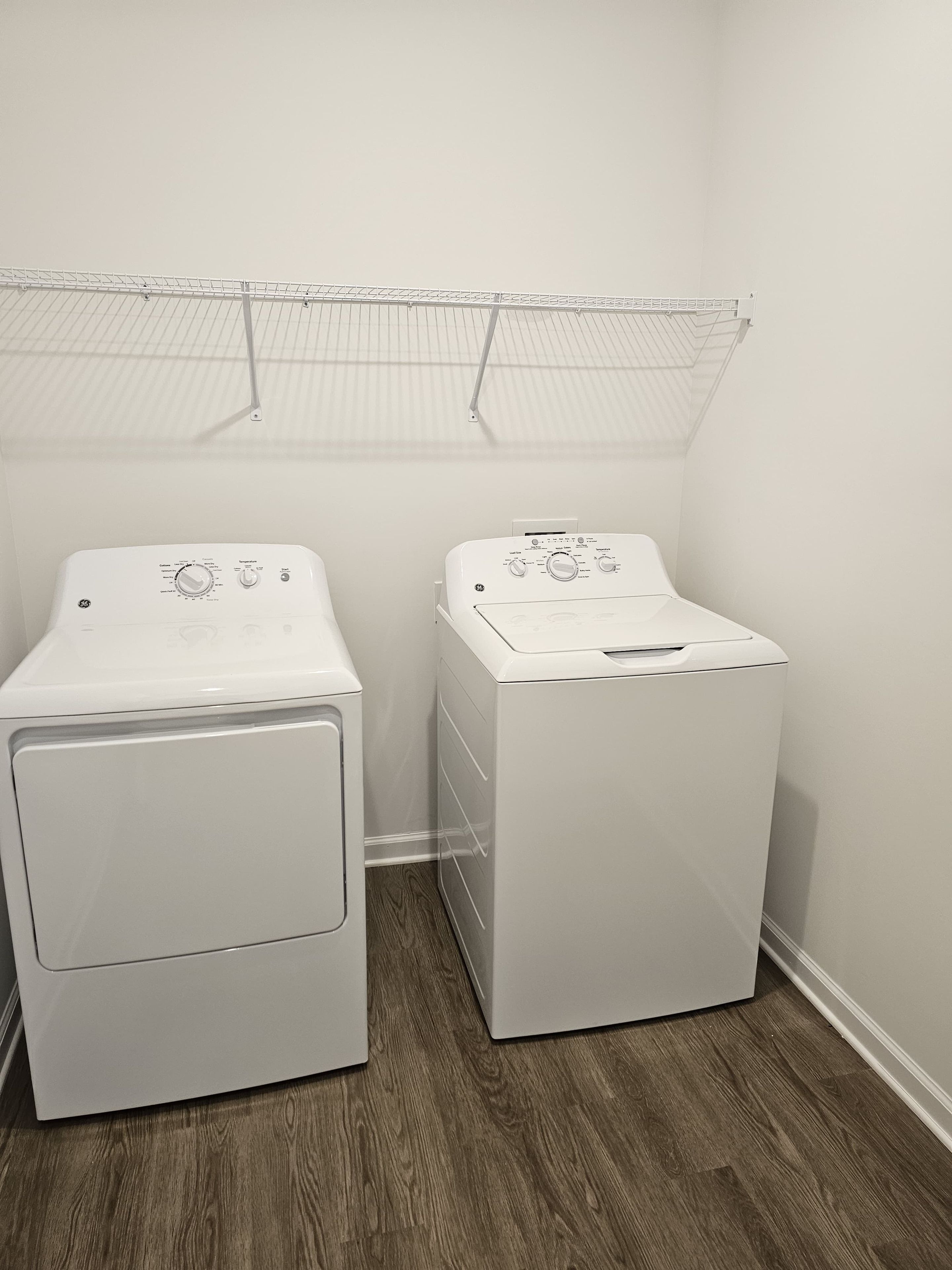 A laundry room with two front-loading washing machines, a metal shelf above, and a wooden floor.