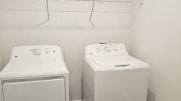 A laundry room with two front-loading washing machines, a metal shelf above, and a wooden floor.