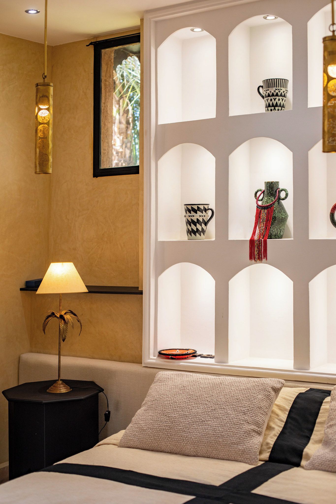 A hotel room with a bed, a side table with a lamp, a window with decorative items, and a wall-mounted shelf.