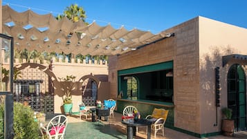Een terras met tafels en stoelen, een pergola voor beschutting en een gebouw met decoratieve muren.
