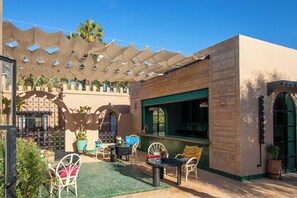 An outdoor seating area with tables and chairs, a pergola with shade, and a building with patterned walls. - Villa Jnane Réhann (Marrakech)