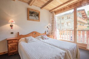 Ein Schlafzimmer mit zwei Betten, Holz-Kopfteilen, Nachttischlampen, einem gerahmten Bild an der Wand und Blick auf ein Steinhaus durch ein großes Fenster.