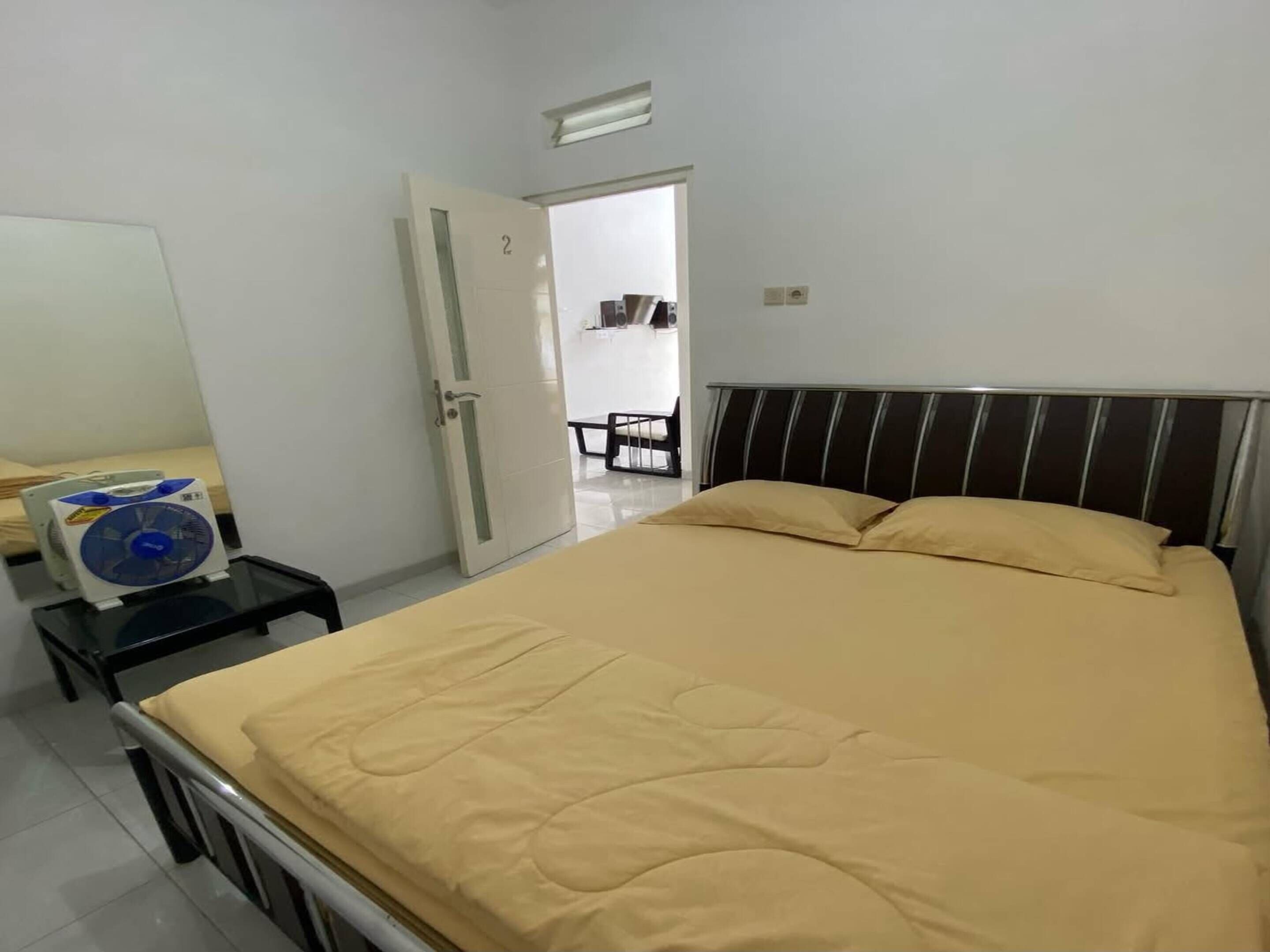 A bedroom with a bed, a bedside table with a fan, a door, and a view of a dining area.