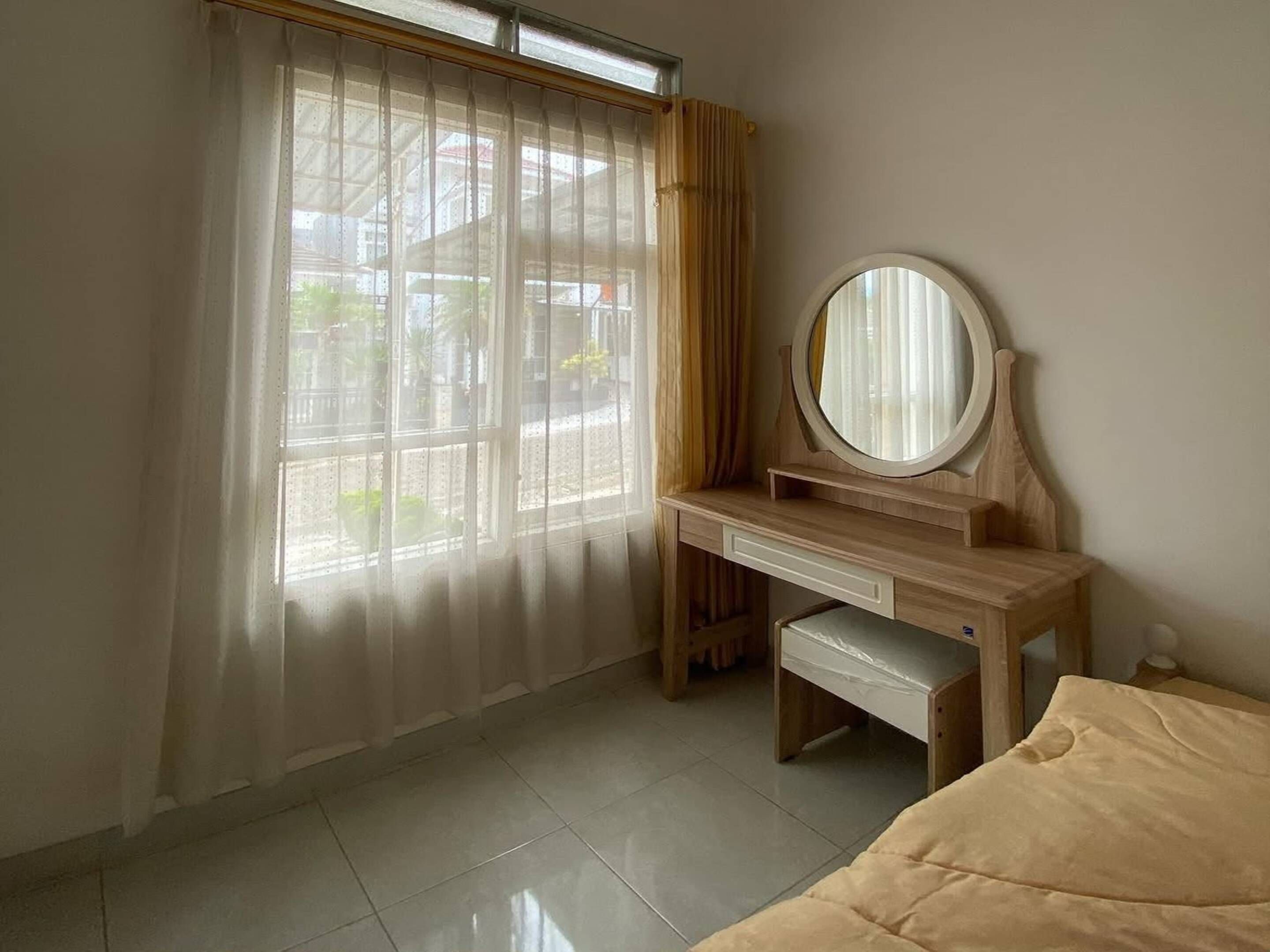 A bedroom with a wooden desk, a round mirror, a bed, and a window with sheer curtains.