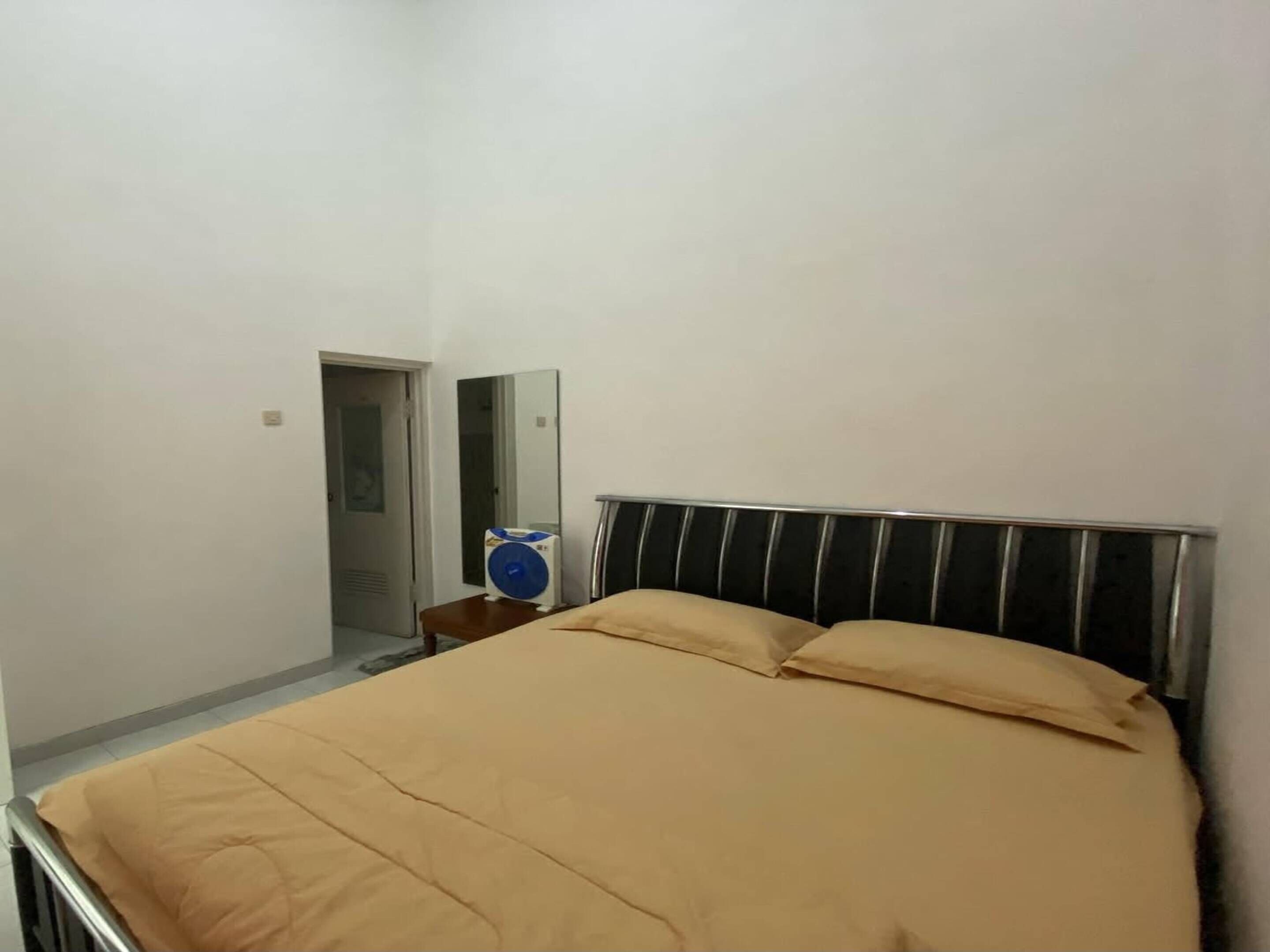 A single bed with a metal frame and beige bedding, a small bedside table with a fan, and a mirror on the wall.
