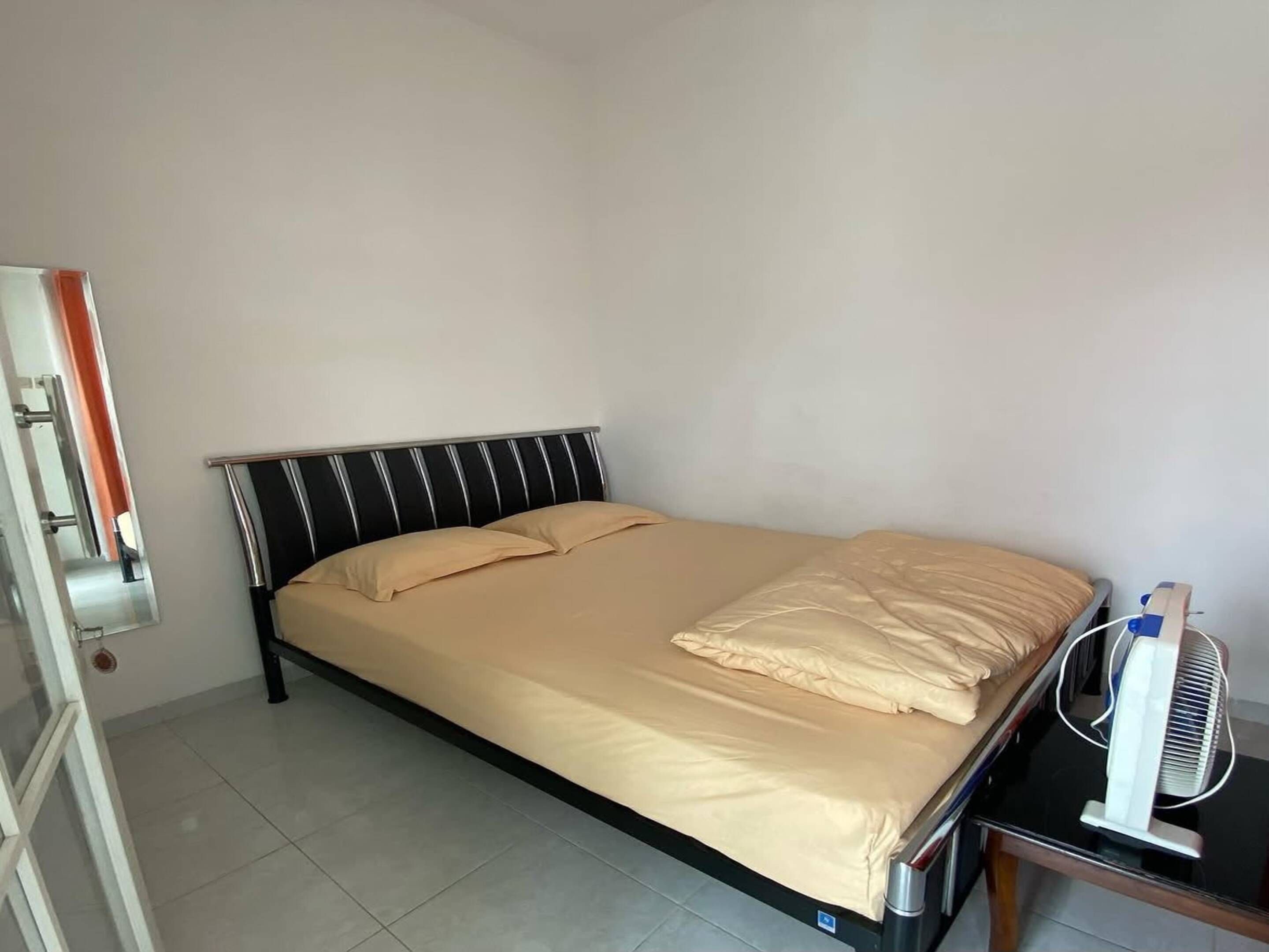 A single bed with a black headboard, a beige mattress, and a white bedside table with a fan.