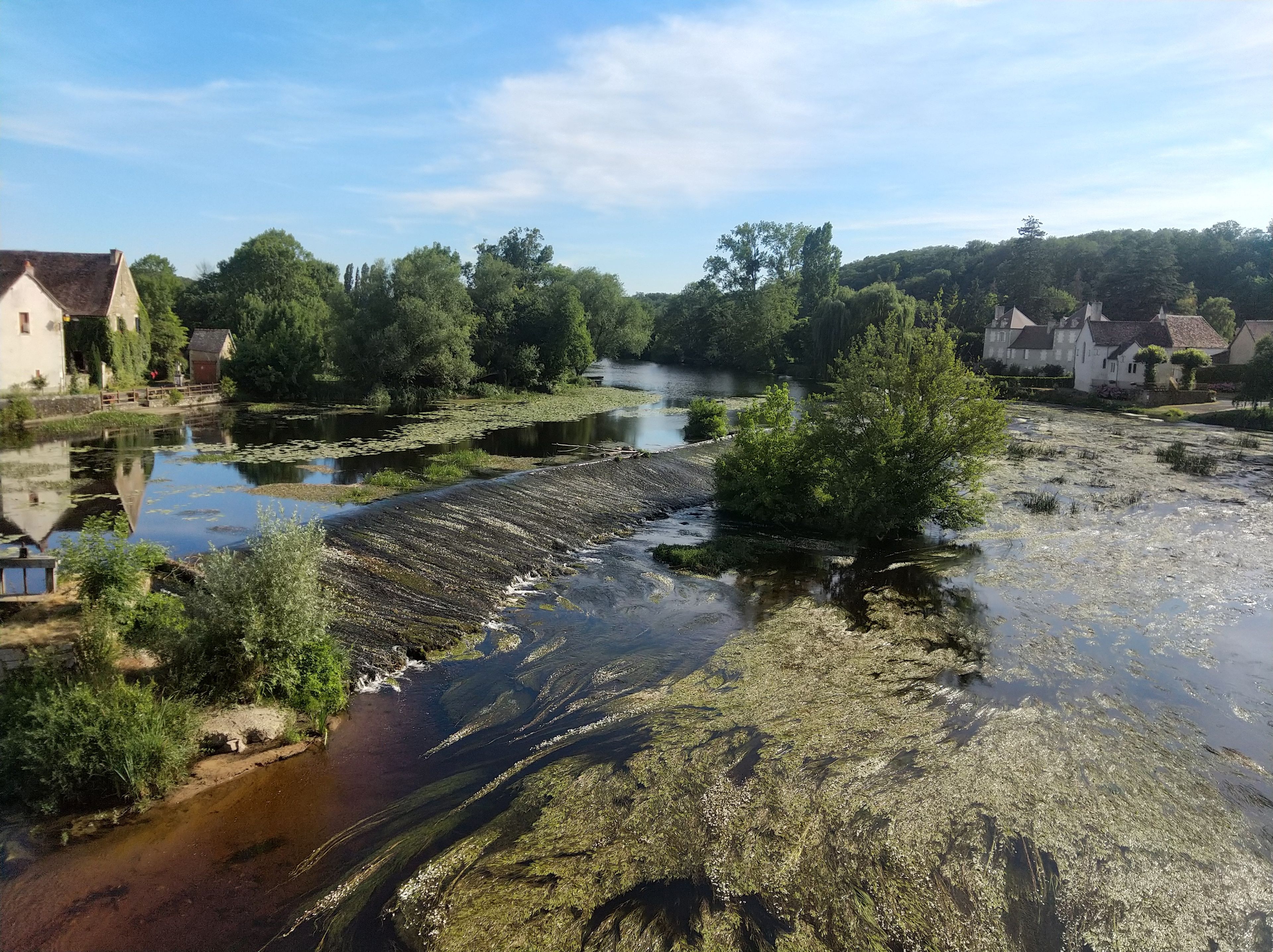 A river with a dam, surrounded by greenery and houses.