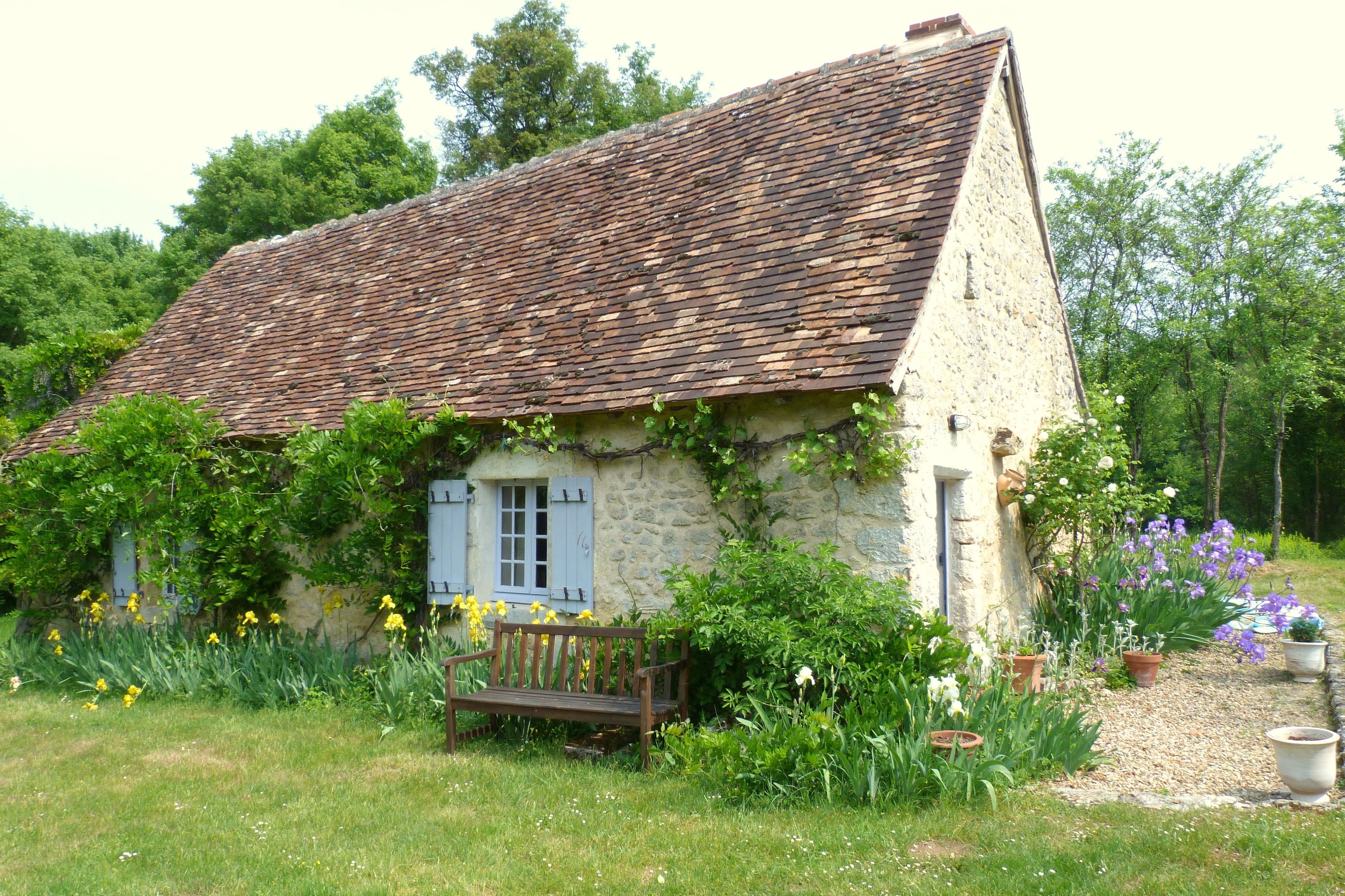 A rustic stone house with a sloped roof, a wooden bench, and a garden with various plants and flowers.