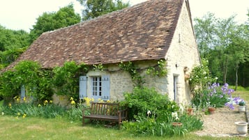 A rustic stone house with a sloped roof, a wooden bench, and a garden with various plants and flowers.