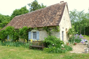 A rustic stone house with a sloped roof, a wooden bench, and a garden with various plants and flowers.