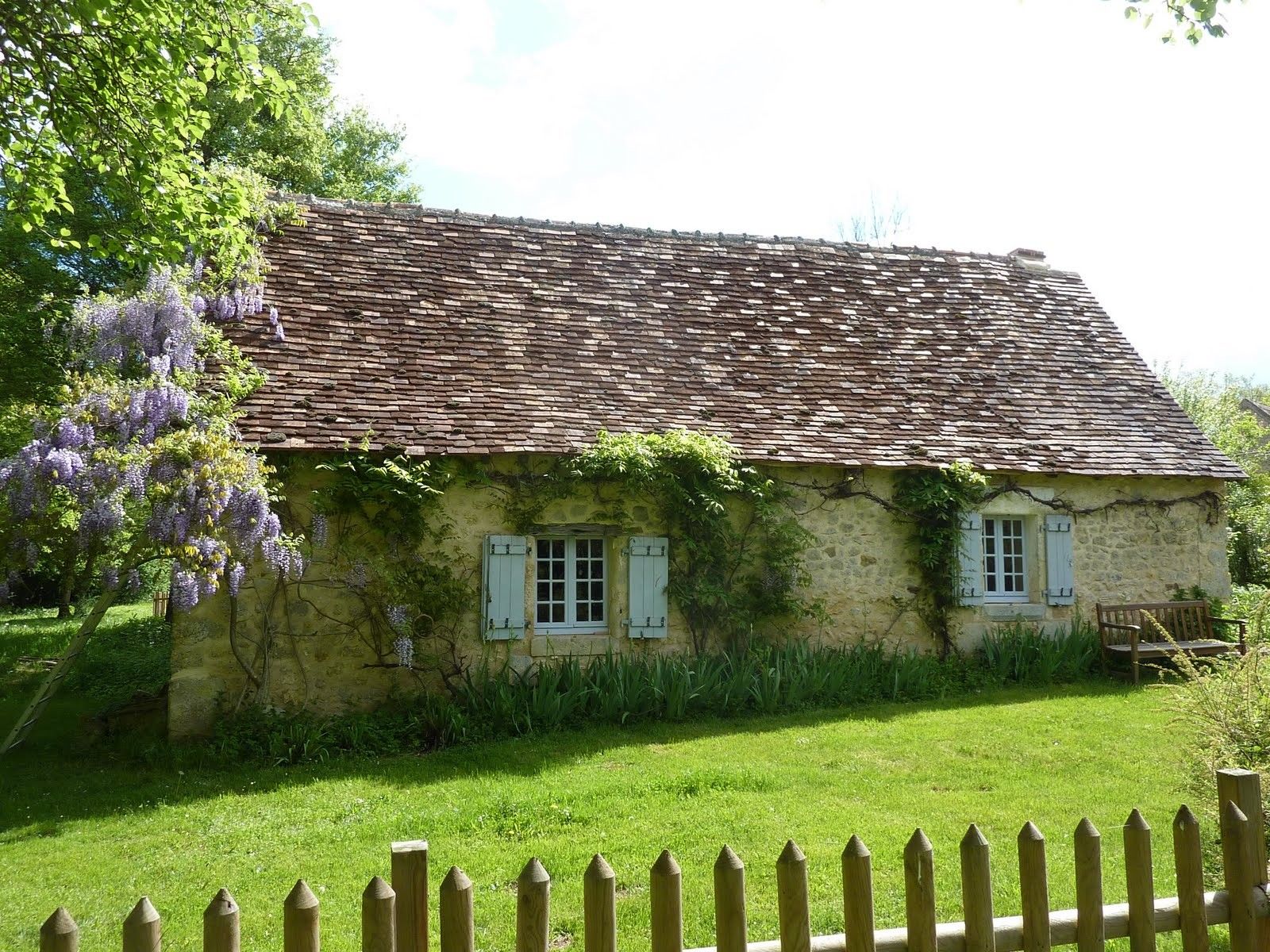 A rustic stone house with a tiled roof, surrounded by a wooden fence and lush greenery.