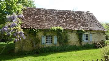 A rustic stone house with a tiled roof, surrounded by a wooden fence and lush greenery.