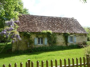 A rustic stone house with a tiled roof, surrounded by a wooden fence and lush greenery.