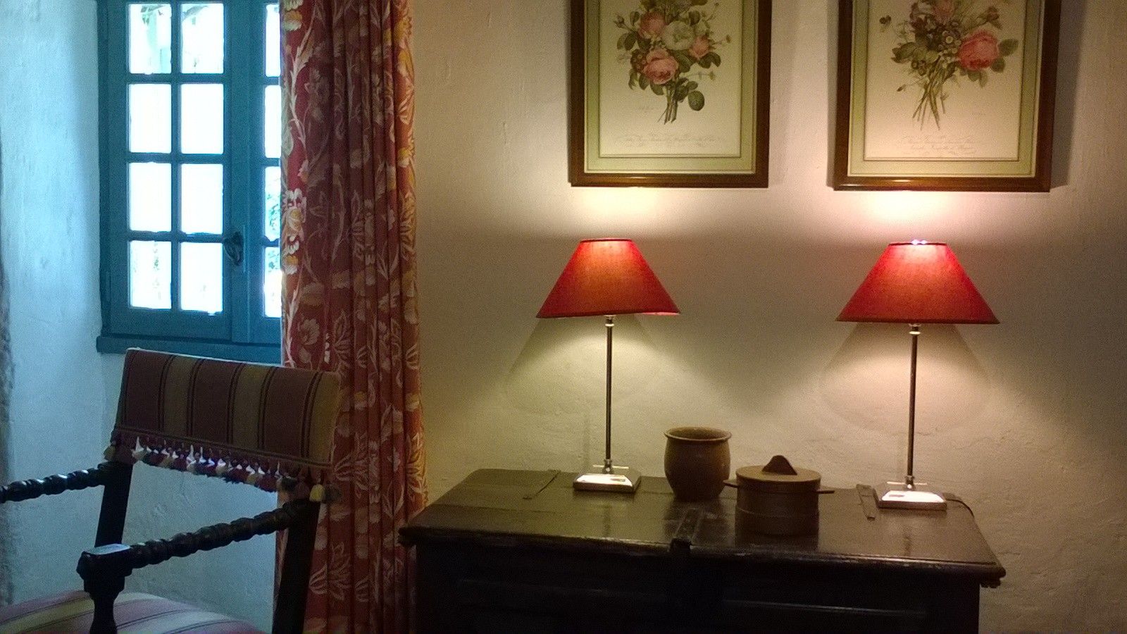 Una habitación con una silla, una mesa con dos lámparas y dos cuadros con motivos florales en la pared.